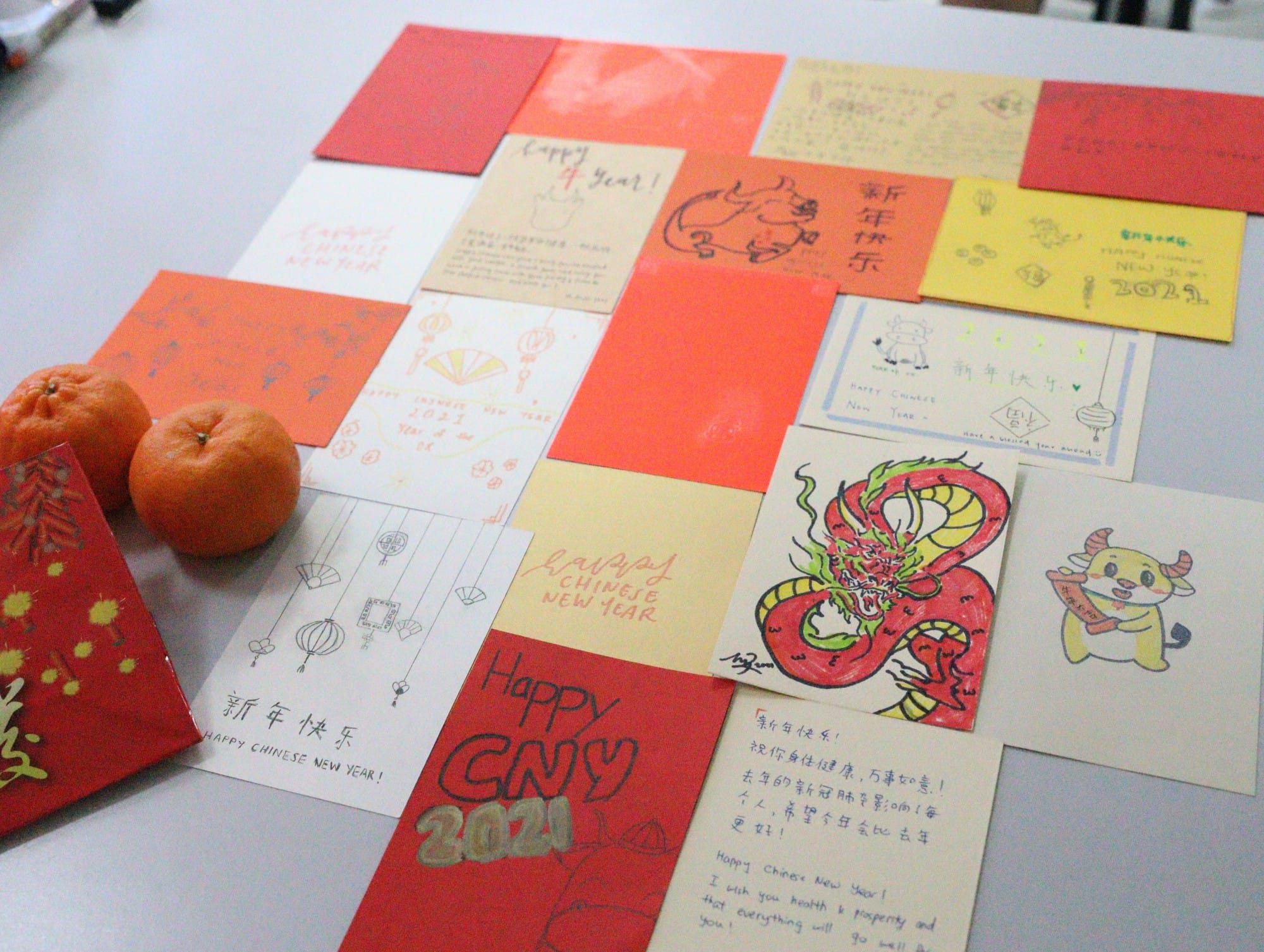 Handmade Chinese New Year cards in red and yellow with drawings and greetings, alongside two mandarin oranges on a table.
